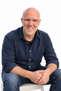 Urs Malke sitzt mit verschränkten Händen mit einem herzlichen Lachen vor der Kamera