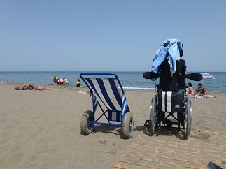 Ein Strandrollstuhl mit dicken Reifen steht im Sand neben einem normalen Handrollstuhl.
